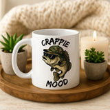 White mug with fish design and 'Crappie Mood' text on a wooden tray with plants and a candle.